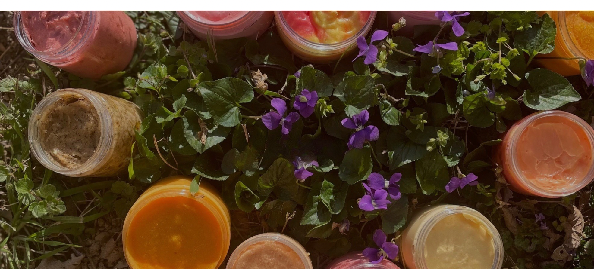 Assorted colorful body scrubs  in plastic cups on a grassy surface with purple flowers.